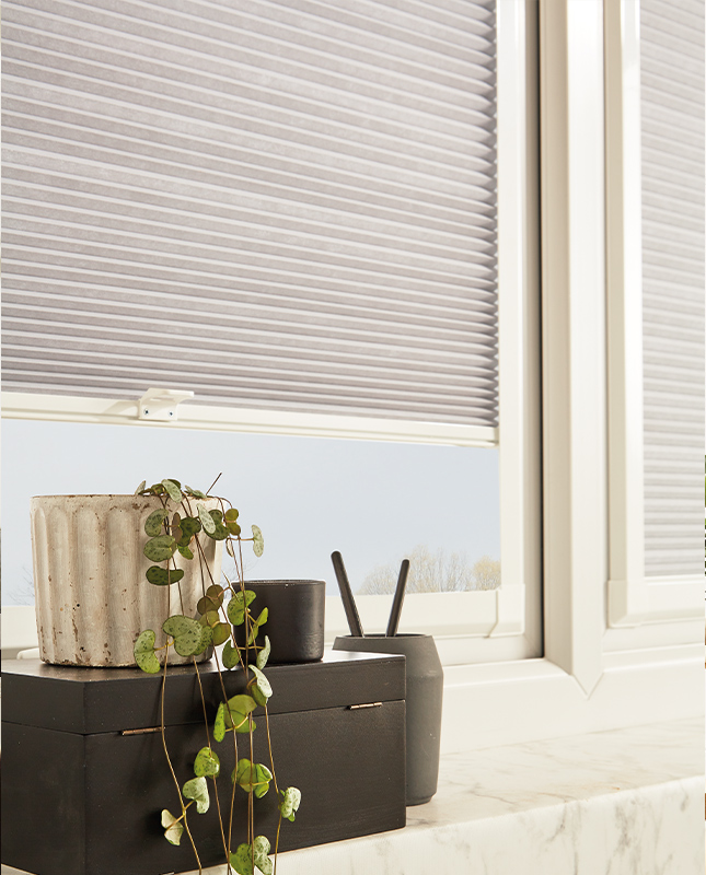 Decorquip grey Perfect Fit pleated blind partially raised above a windowsill with a trailing plant