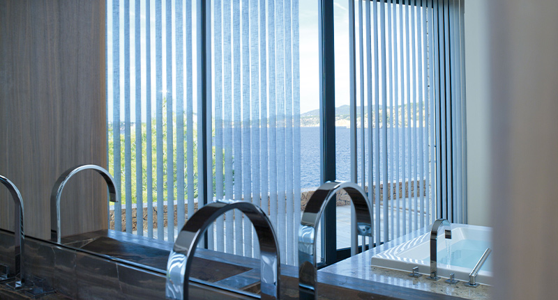 Decorquip white vertical blinds covering floor-to-ceiling windows in a modern bathroom with sea views